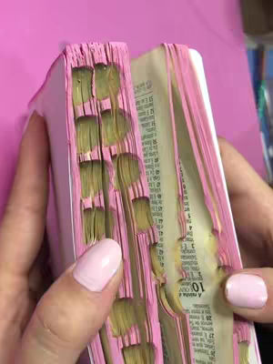A close-up shot displays a well-worn Bible, open to a page with visible text. The edges of the pages are dyed pink, and the binding appears aged. A hand with pink nail polish holds the book open, emphasizing the well-loved condition of the Bible.