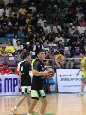 The image shows a basketball game in progress. A man in a black athletic shirt and green shorts is holding a basketball and walking on the court. Behind him, another player in a black jersey with the number 10 is walking. In the background, a crowd of people is seated in the stands. The scene is brightly lit, suggesting an indoor arena or stadium setting.