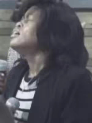A woman with black hair is looking upwards with her mouth open. She is wearing a black jacket over a black and white striped shirt. A microphone is in the shot.