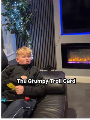 A young boy sits on a black couch in front of a dark gray paneled wall and a lit electric fireplace and a tv. He holds a yellow card in his right hand and watches the card with a surprised facial expression. The text overlay says, "The Grumpy Troll Card."