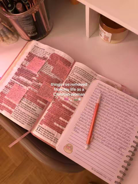 An overhead shot featuring an open Bible and a spiral notebook on a white surface. The Bible has pink sticky notes highlighting passages. A pencil rests on the notebook, which has handwritten notes. There are also makeup brushes, a jar, and a shelf in the background which create a cozy atmosphere.