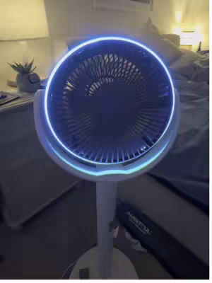 The image shows a white fan with a glowing blue light ring around the fan blades. The fan is on a stand, and in the background, there is a bed, a bedside table with a plant, and a lamp. The overall composition is well-lit, with a focus on the fan.