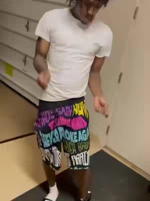 A man is dancing in a room. He is wearing a white t-shirt and colorful shorts that say "Never Broke Again".