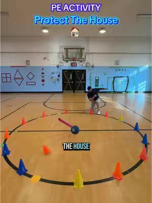 A low-angle shot captures a gymnasium with a wooden floor marked with lines for a game of 'Protect the House.' Cones of various colors ring the center of a circle, around which a player, the main subject, is poised, ready to throw a ball. The background shows the gym's infrastructure: a basketball hoop, a wall with shapes and the word 'GOAL', and doors. Text overlays, including 'PE ACTIVITY,' 'Protect The House,' and 'THE HOUSE,' frame the scene as playful and instructional.