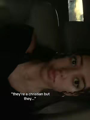 A selfie-style video cover. The creator, a person with dark hair, is seen from the chest up, slightly off-center. The lighting is dim, creating shadows and emphasizing the face. Written text overlays the image, in white font, saying "they're a christian but they...". The background is dark and indistinct.