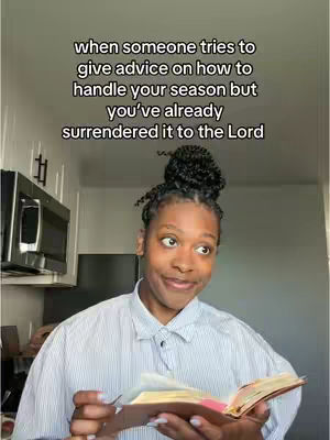 A woman with a bun hairstyle is holding a book, likely a Bible, and looking to the side with a slight smile. The background consists of a kitchen setting with white cabinets and a microwave.