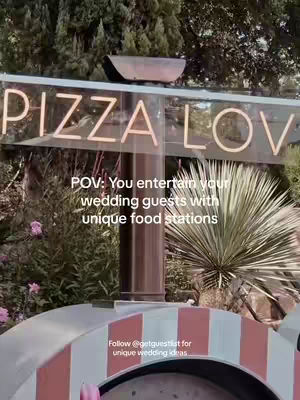 The video shows a vertical shot of a pizza restaurant with a sign that reads 'PIZZA LOV'. Below the sign there is written text that states, 'POV: You entertain your wedding guests with unique food stations.' The bottom portion of photo shows the roof of the building which has a pink and white striped pattern. The background includes greenery such as trees and shrubs.