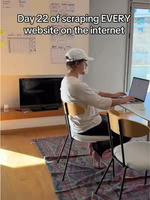 A man sits at a desk in a well-lit room, working on a laptop. Behind him is a wall with some papers and a TV screen.