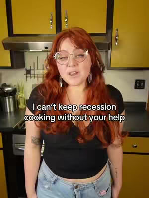 A woman with red hair, wearing glasses and a black top, stands in a kitchen. Text overlayed on the image says "I can't keep recession cooking without your help". The woman is looking directly at the camera, the background includes yellow cabinets and kitchen appliances.