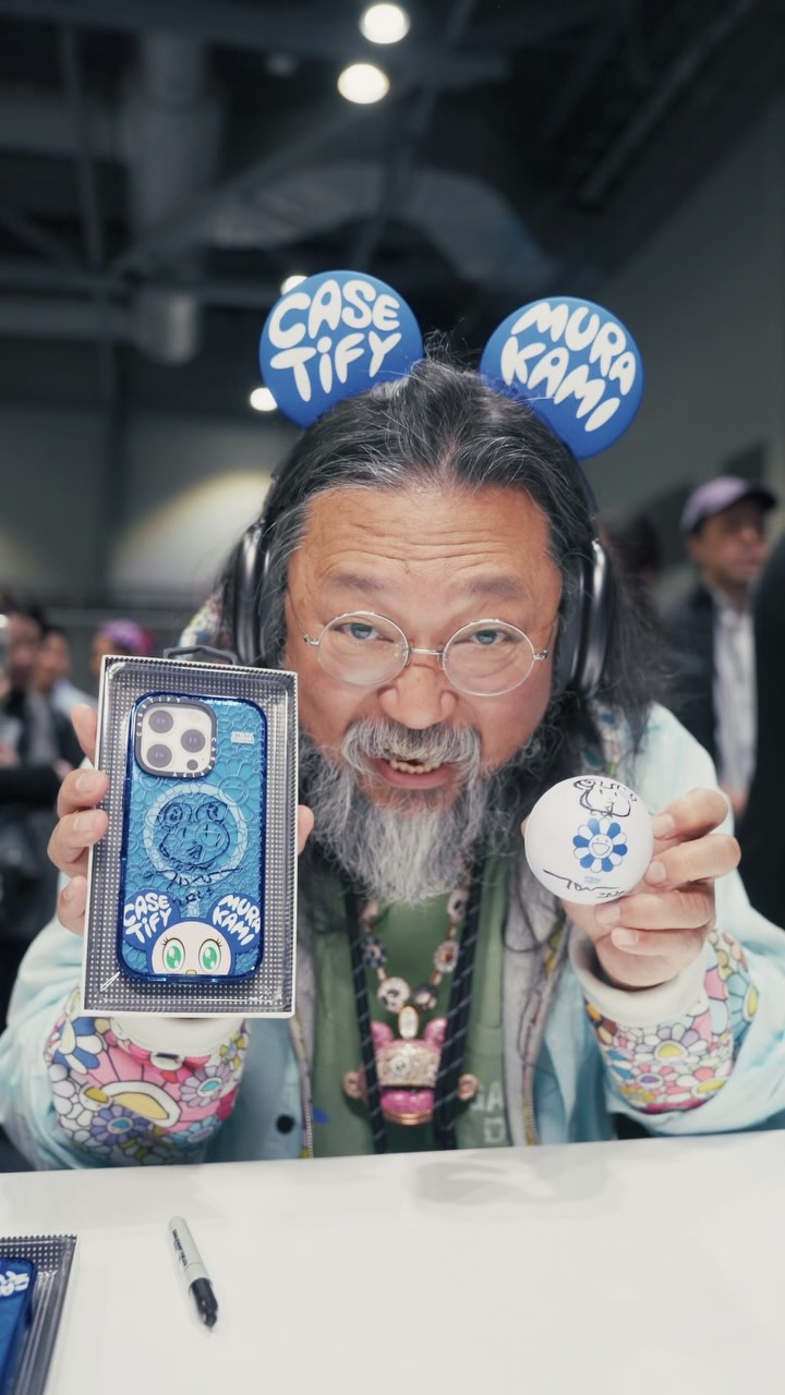 A man with long gray hair and glasses holds up a phone case and ball, both featuring colorful designs, smiling at the camera. He wears a headset, a light blue jacket, and is surrounded by what appears to be a convention setting with other people in the background.