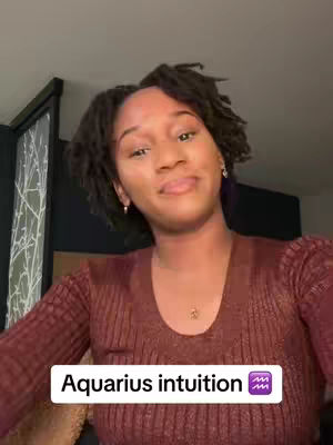 A woman with locs is smiling and looking slightly to the side. She wears a maroon top and small gold earrings and necklace. Text overlayed on the bottom reads, "Aquarius intuition". The background is a dark room.