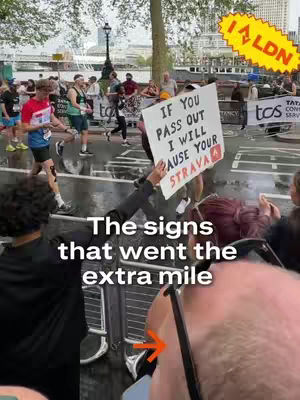 A photo taken from a lower angle showing a marathon race in progress. In the foreground, people are visible, including the back of a person's head and a person holding a sign. The sign displays a humorous message concerning Strava. Runners on the race are showing, the sky is overcast. The image has a mix of natural and artificial lighting with a muted color palette.