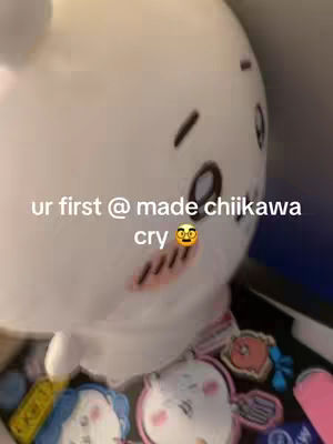 The image shows a plush Chiikawa toy from the side, slightly out of focus. Text overlayed reads, "ur first @ made chiikawa cry 🥺" with an emoji. The background is blurred, suggesting an indoor setting with other toys or items partially visible.