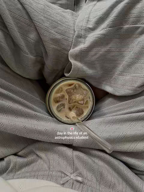 An overhead shot shows a cup of coffee on a grey, textured blanket. The coffee is light brown, with a star-shaped ice cube floating on the surface. A clear straw extends from the cup. The blanket is folded in a way that the cup is nestled within. A bow is tied at the bottom part of the blanket.