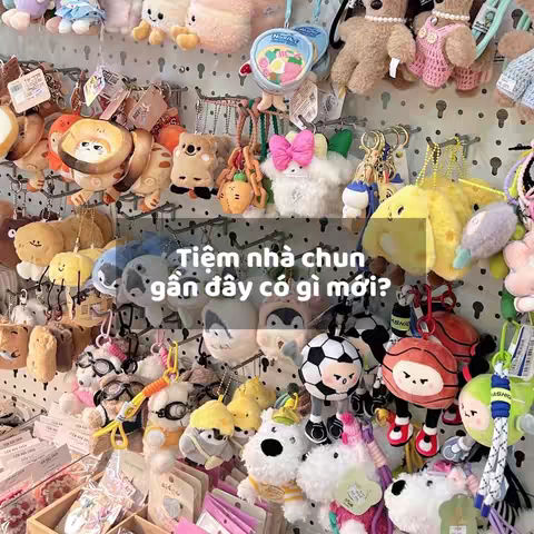 A display of various keychains is shown, hanging densely in front of a pegboard-style backdrop. The keychains feature a range of plush characters and objects. The text overlay is in Vietnamese.