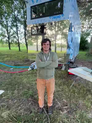 A man stands with arms crossed in front of a partially constructed building. The building is made of reflective material, and its frame is visible. Various tools and materials are scattered on the ground, and the background includes trees and a grassy area.