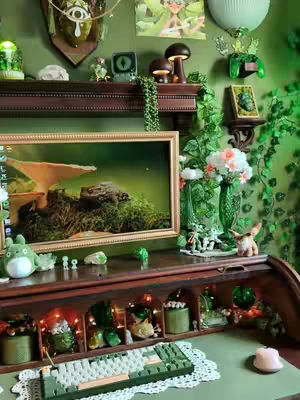 An aesthetically arranged desk with a vintage rolltop design. The desk is laden with a computer, a green-themed keyboard, and various cozy decorations. An animated desktop background is visible. Soft lighting and a blend of natural and artificial elements create an inviting atmosphere.