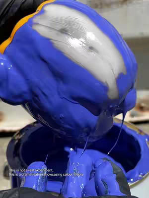 A close-up shot of a container filled with a vibrant blue, viscous liquid, with a patch of white or silvery liquid in the center. The blue liquid appears to be spilling over its rim. Paint drips are visible. Black gloves are present in the scene, holding the container.