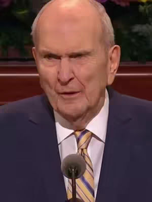 The image shows a close-up of a man of senior age, likely in his 80s or 90s, with a neatly trimmed hairline and thin, styled fair hair. He has a strong nose, deep-set eyes, and is mid-speech with his mouth slightly open, appearing to be addressing an audience. He wears a dark navy suit over a white collared shirt and a tie with yellow and blue diagonal stripes. A microphone is positioned in front of him at the bottom of the frame, suggesting a formal setting such as a conference or presentation.