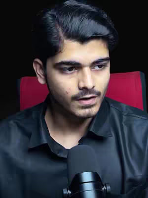 A medium shot features a young man with dark hair, a mustache, and a beard, gazing slightly to the side. He wears a black shirt, with a microphone positioned in front of him. The background is dark, with a red chair visible behind him.