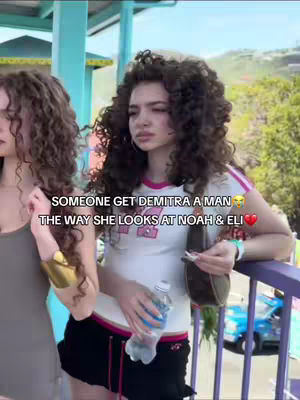 The cover image shows a medium shot of three young women in an outdoor setting. The focus is on the central figure, who is looking off to the side with a slightly wistful expression. She is wearing a white t-shirt with pink accents and black shorts. Next to her is another woman with long curly hair. In the background, there is a partially visible building, trees, and a blue car, suggesting an urban or recreational area. The overall composition uses a natural color palette enhanced with added text.