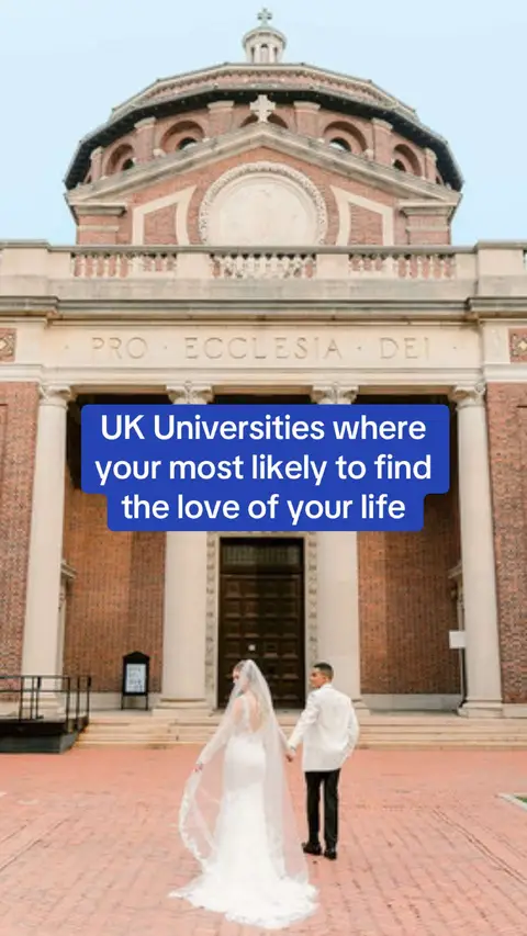 A wedding couple, a bride and groom, are walking away toward the entrance of a brick building. The bride is in a white dress with a long veil, and the groom is wearing a white blazer and black pants. The building has a large arched entrance with a sign above it.