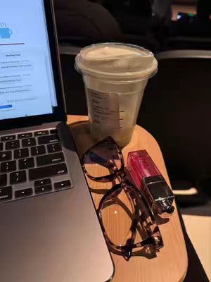 A laptop on a wooden surface with a clear iced coffee cup, tortoise shell glasses, and a pink lip gloss.