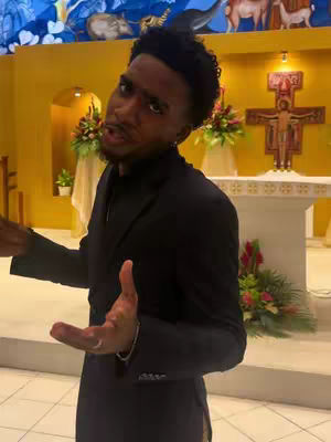A man in a black suit is gesturing with both hands, speaking in what appears to be a church. Behind him, a crucifix and floral arrangements are visible. The background features architectural details and religious artwork. The lighting is relatively bright, illuminating the setting.