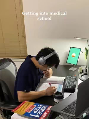 A well-lit photo features a young man at a desk, engrossed in studying. He wears large gray headphones and glasses. A computer, tablet, and books are strewn across the desk. Text overlayed on the image reads, “Getting into medical school”.