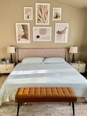 A cozy bedroom scene featuring a bed with light blue bedding. The bed is centered, with a brown bench at its foot. On the wall above the bed, there are several framed artworks.