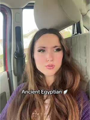A selfie-style shot taken from inside what appears to be a car. A young woman with long brown hair and visible makeup is the primary focus, set against a backdrop of car interior elements and a window with a hint of an outdoor scene. The color palette is dominated by neutral tones from the car's interior, complemented by the woman's warmer skin tone and a purple sweater.