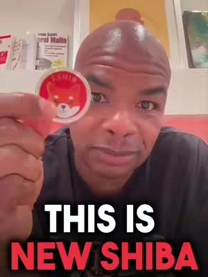 A man with a shaved head holds up a Shiba Inu coin, with a text overlay at the bottom reading 'THIS IS NEW SHIBA'. He is positioned in front of a background with framed pictures and text on display, presenting a focus on cryptocurrency.