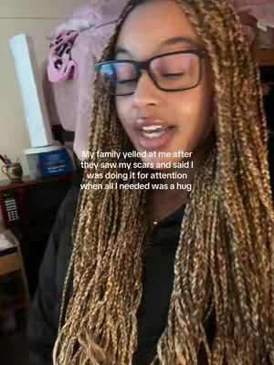 A young woman with long, braided hair and glasses is the central focus. The background is blurred, but appears to be a room, with a light-colored object and other items partially visible. Text is overlaid on the image.