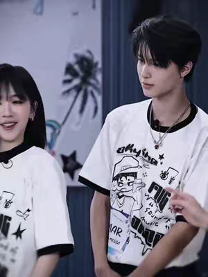 The cover image shows two young people standing side-by-side in a brightly lit setting, likely indoors. Both wear white t-shirts. The person on the right has their right arm bent, possibly holding or gesturing with their hand. The t-shirt on the right has a unique design with images and text in a cartoon style, while the left one seems to have a simple logo. The background is blurred but suggests an event or studio setting. Soft lighting and a neutral color palette dominate the scene.