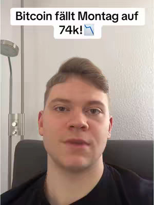 A medium shot shows a man with short, light brown hair, wearing a black t-shirt, seated in a chair. Above his head, a text overlay states 'Bitcoin fällt Montag auf 74k!' with a downward trend arrow. The background is a plain wall with a silver lamp.