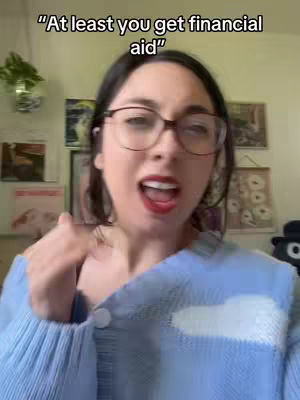 A woman with dark hair and glasses is looking at the camera, mouth open as if talking or singing. She's wearing a light blue sweater. The background includes framed pictures and a hint of greenery.