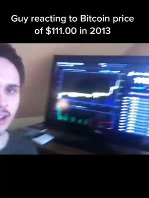 A man is shown in the video, with a portion of his face visible on the left-hand side. Behind him, a computer monitor displays a financial graph. The overlaying text at the top reads, 'Guy reacting to Bitcoin price of $111.00 in 2013.'