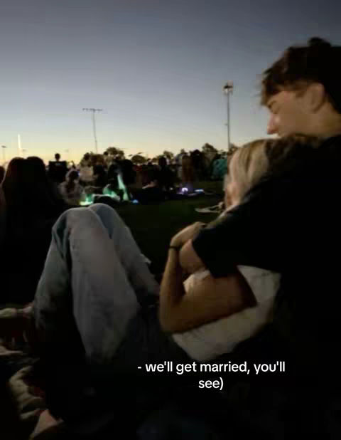 A couple sits together on a grassy area at dusk, with people and lights in the background. The man is embracing the woman, and a text overlay is at the bottom.