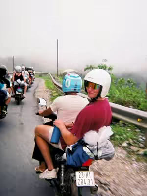 A photo taken from behind of two people riding a motorbike on a road. The person in the front is wearing a white helmet with a clear visor, sunglasses and a red shirt. The person in the back is wearing a light blue helmet and white shirt. The road is wet and there is greenery on either side. Other riders are also riding down the road with them.