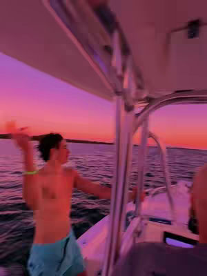 A person on a boat, with an orange and purple sunset in the background. The person is shirtless with blue shorts and is moving their arms. The composition gives a sense of motion and freedom.