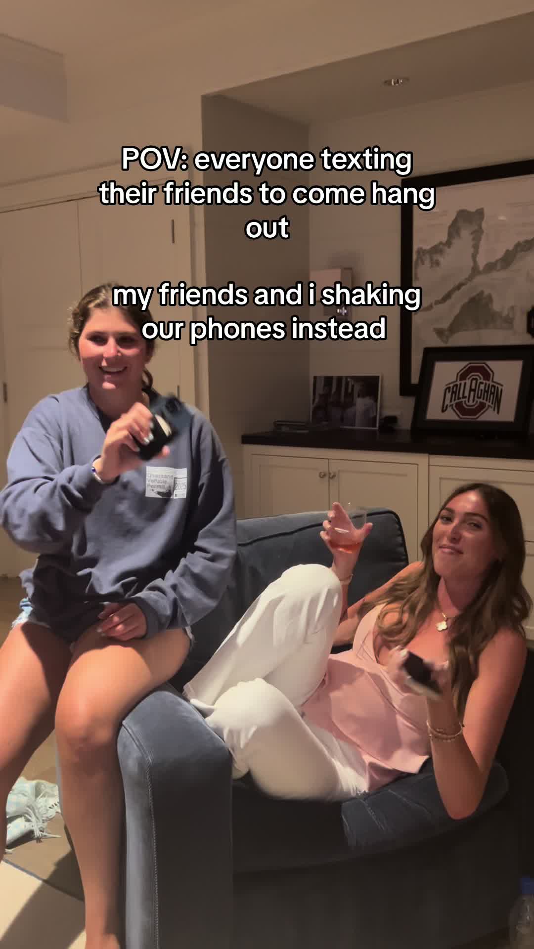The cover image captures a moment inside a well-lit living space. Two young women are the primary subjects: one is seated in a cozy armchair to the left, holding up a black phone and smiling. The other is seated on the armrest. The background features a partially visible wall with decor and a glimpse of what look like a study or office space. The overall composition suggests a casual, indoor setting.