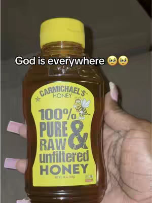 A hand holding a bottle of Carmichael's Honey. The bottle is mostly transparent with a yellow cap and a yellow label on the front. On the label there is text that says: 'Carmichael's Honey', '100% PURE & RAW unfiltered HONEY'. There is a drawing of a bee on the label. The hand holding the bottle has long nails painted pink.