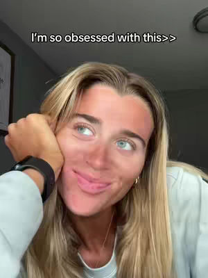 A close-up shot of a young woman with long blonde hair. She is touching her cheek with her hand and looking toward the side, with her lips slightly parted in a subtle smile. The background is blurry and out of focus.
