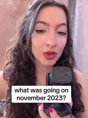 A woman with long, wavy dark hair is centered in the frame, gazing directly at the camera. She's holding a black microphone and wearing a dark top. Overlaid on this image is a white rectangle containing the text 'what was going on november 2023?'. The background is blurred and warm-toned, suggesting an interior setting.
