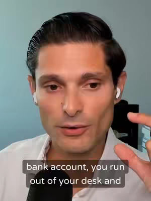 A man with short, slicked-back dark hair, wearing a white shirt and white earbuds, is centered in the frame. His expression is serious, with engaged eyes. A dark microphone is at the bottom of the frame. Below the man, overlayed text, in a black box, reads, 'bank account, you run out of your desk and'.