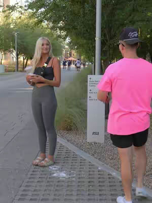 A woman with blonde hair is standing on a sidewalk looking toward a man in a pink shirt. The man is turned away from the camera. They seem to be in a city park.