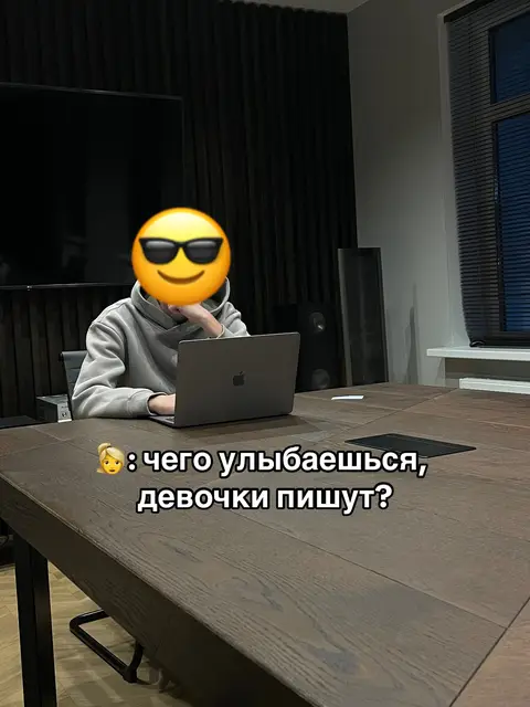 The image shows a person seated at a dark wooden table in a modern, dimly lit room. The person's face is obscured by a sunglasses emoji, wearing a gray hoodie and using a laptop. Text overlays the image, suggesting a humorous interaction or a potential romantic interest based on the caption. The background includes black vertical paneling, a large TV, and blinds near a window, indicating a comfortable and private setting.