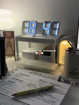 A well-lit desk setup featuring a digital clock displaying '88:44'. A notebook with handwritten notes and a pen lie in the foreground. A Snoopy-themed mug and lamp are visible.