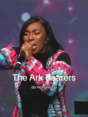 A woman with dark skin is on a stage, holding a microphone and speaking passionately. She wears a black top under a colorful patterned jacket. The background is a blurred gradient of purple and blue with stage lights. Text overlays in white are at the bottom, reading: 'The Ark Bearers', and below that, 'do not give up'.