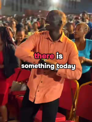 A man in an orange shirt dances joyfully in front of a crowd. The text overlay says "there is something today".